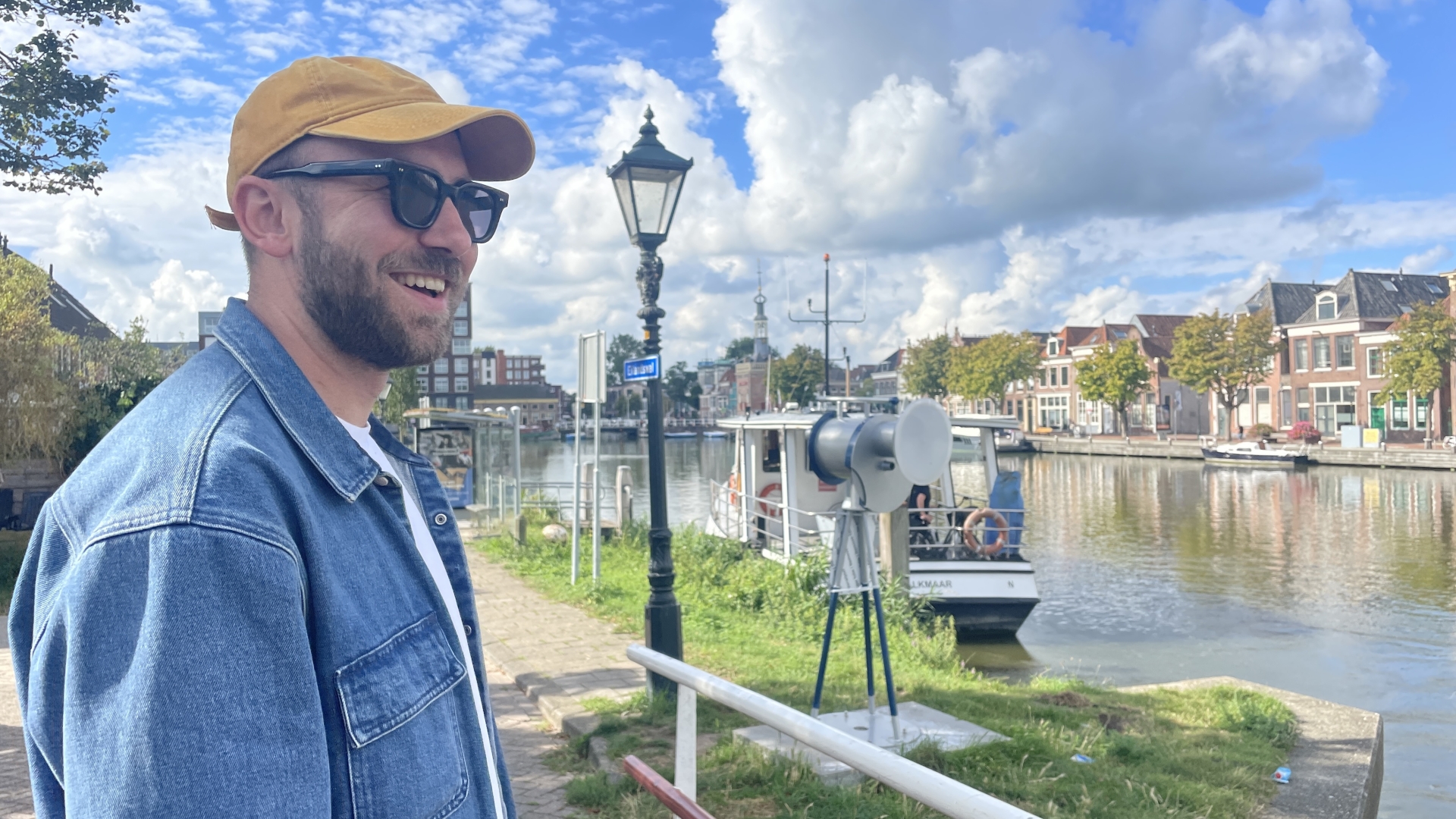 Man met gele pet en zonnebril staat bij een gracht met boten en historische huizen in de achtergrond.