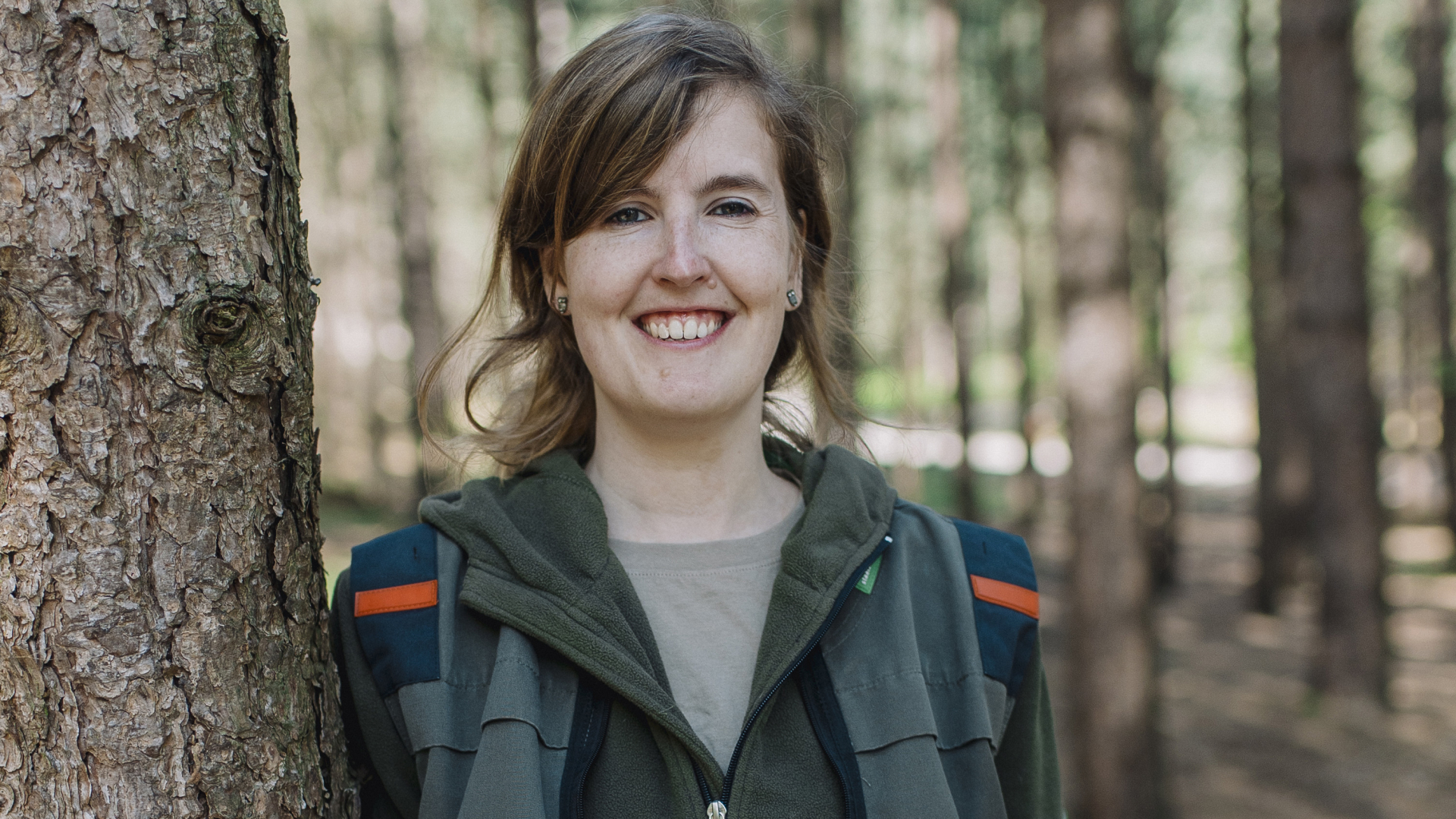 Vrouw leunend tegen een boom in een bos lachend in de camera.