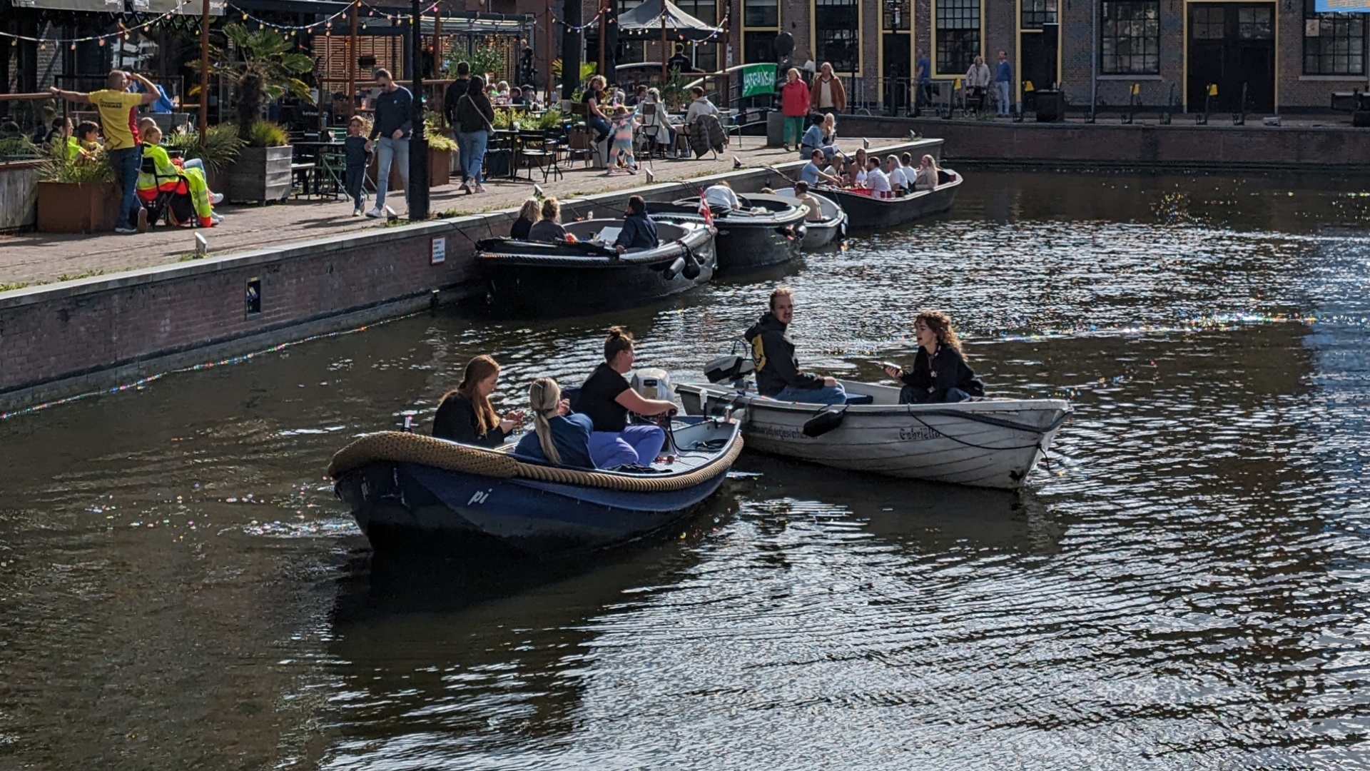 Mensen varen in boten op een gracht omringd door gebouwen en caféterrassen.