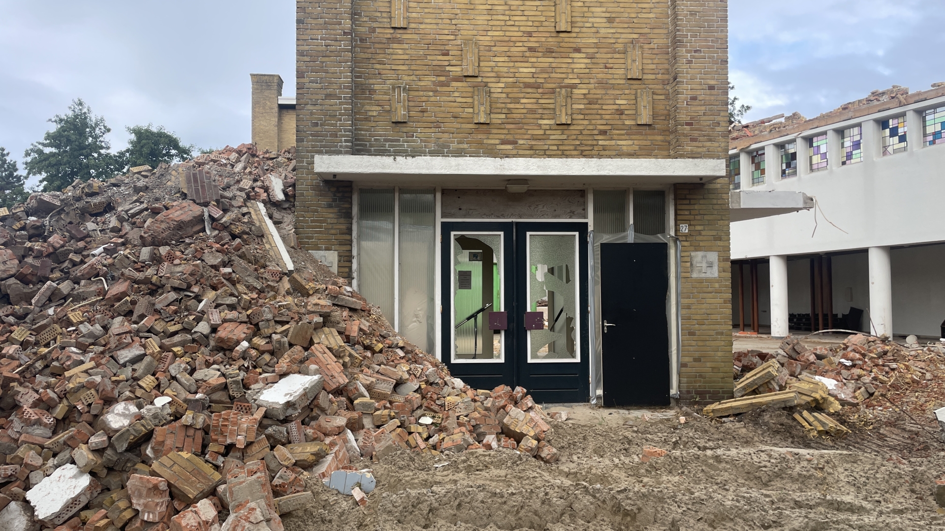 Gezicht op een gedeeltelijk gesloopte bakstenen muur naast een gebouw met gebroken deuren en ramen.