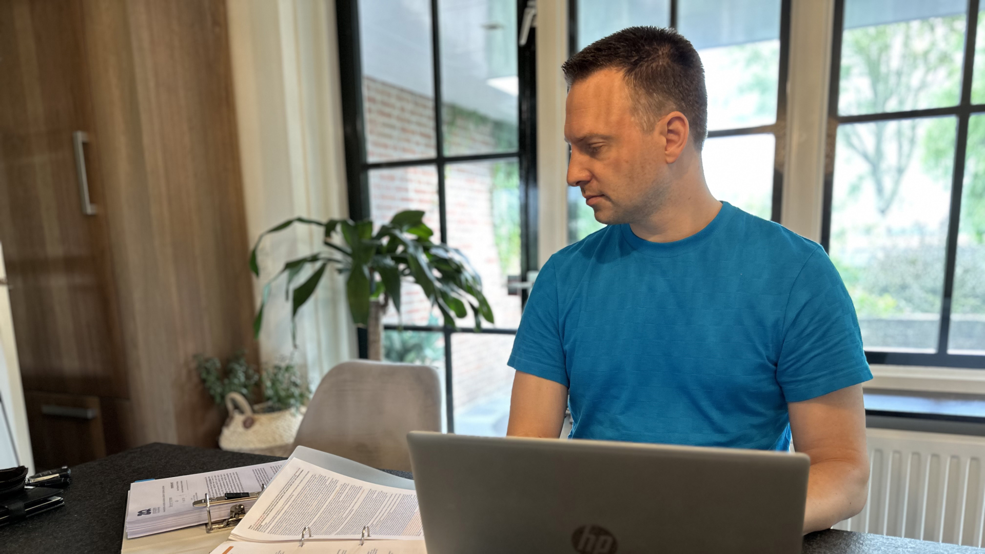 Man in blauwe trui werkt aan laptop met documenten op tafel in lichte kamer.