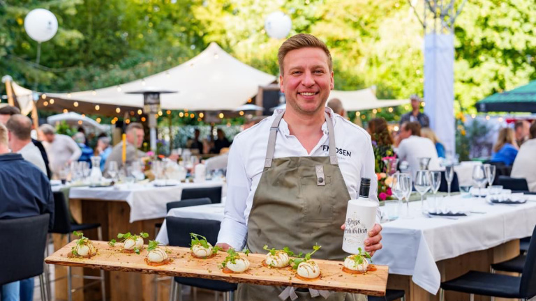 Man in schort serveert een houten plank met kleine gerechten tijdens een buitenfeest.