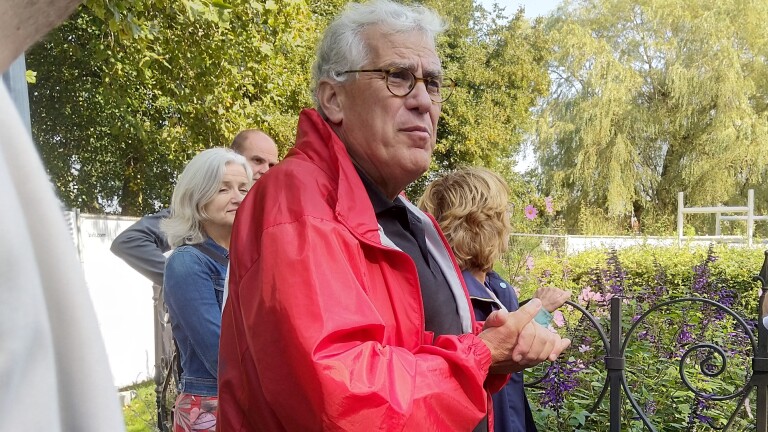 Een oudere man met een grijze haren en een rode jas staat buiten met een groep mensen in een tuin.