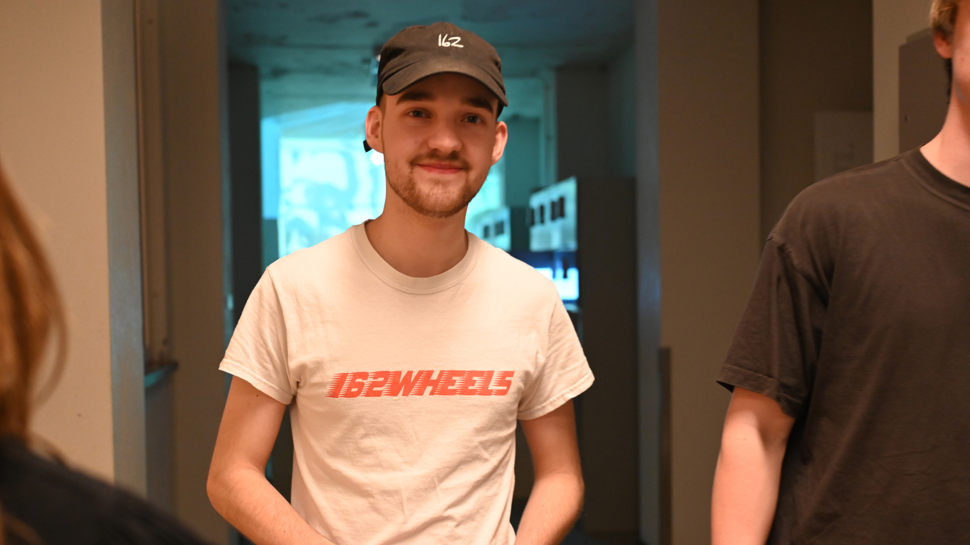 Jonge man met een zwarte pet en een wit T-shirt met rode tekst "152WHEELS" glimlacht naar de camera.