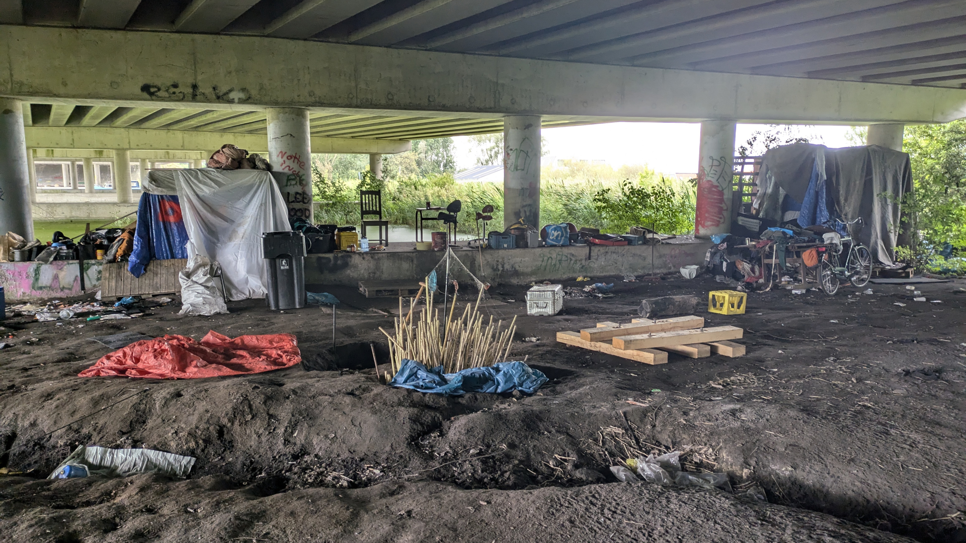 Onderkant van een brug met een geïmproviseerde woonruimte, inclusief fietsen, meubels, tenten en verschillende bezittingen.