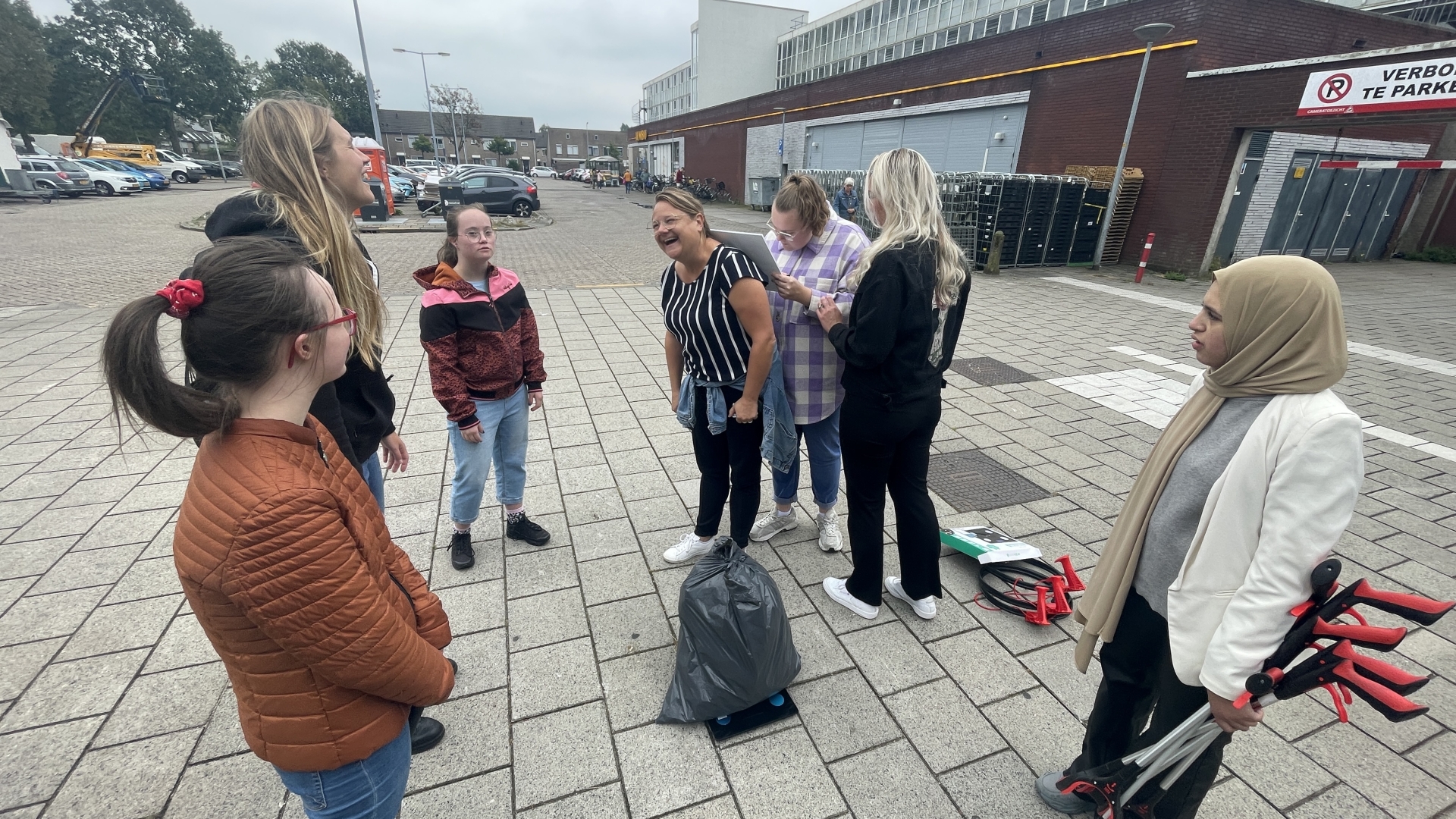 Een groep mensen staat buiten in een stedelijke omgeving, lachend en met elkaar pratend, met afvalknijpers en een vuilniszak op de grond.