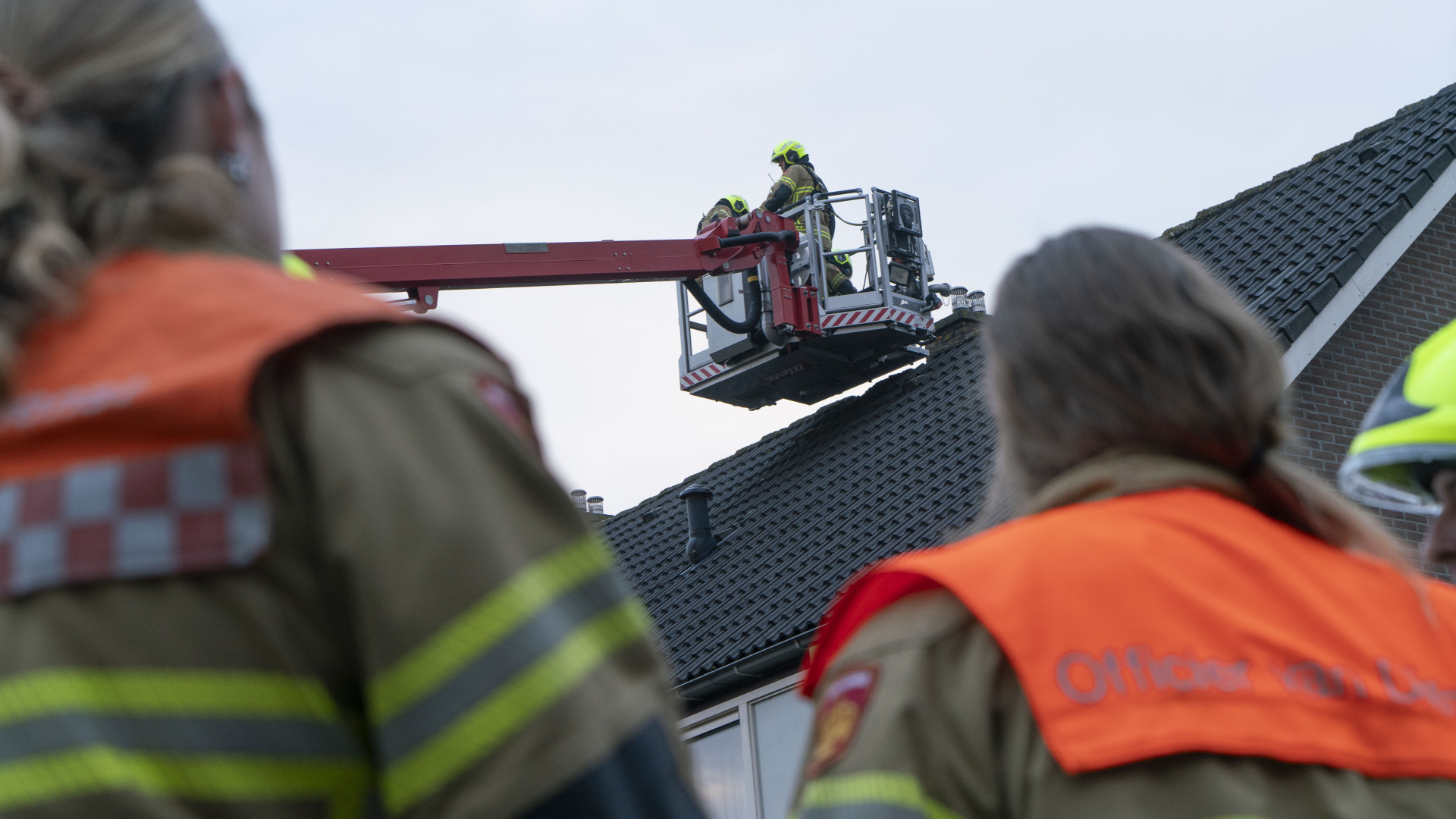 Brandweerlieden werken op een ladderwagen bij een dak, terwijl collega's toekijken.