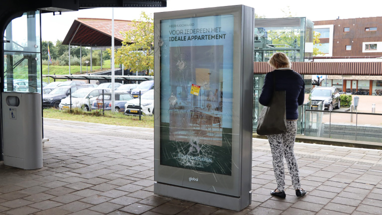 Een persoon staat voor een bushalte met een beschadigd reclamebord dat een advertentie voor appartementen in Heerhugowaard toont.