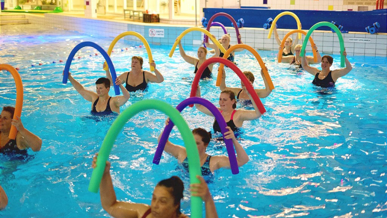 Mensen doen aan wateraerobics in een zwembad, gebruikmakend van kleurrijke schuimrubberen buizen.