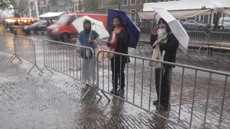 Drie mensen staan buiten in de regen onder paraplu's achter een hek; één persoon zwaait.