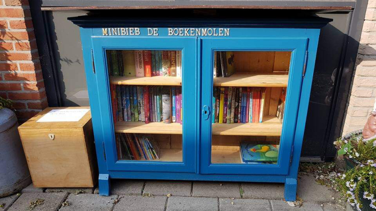 Blauwe minibibliotheek met dubbele glazen deuren, gevuld met boeken, en tekst "MINIBIEB DE BOEKENMOLEN" bovenop.