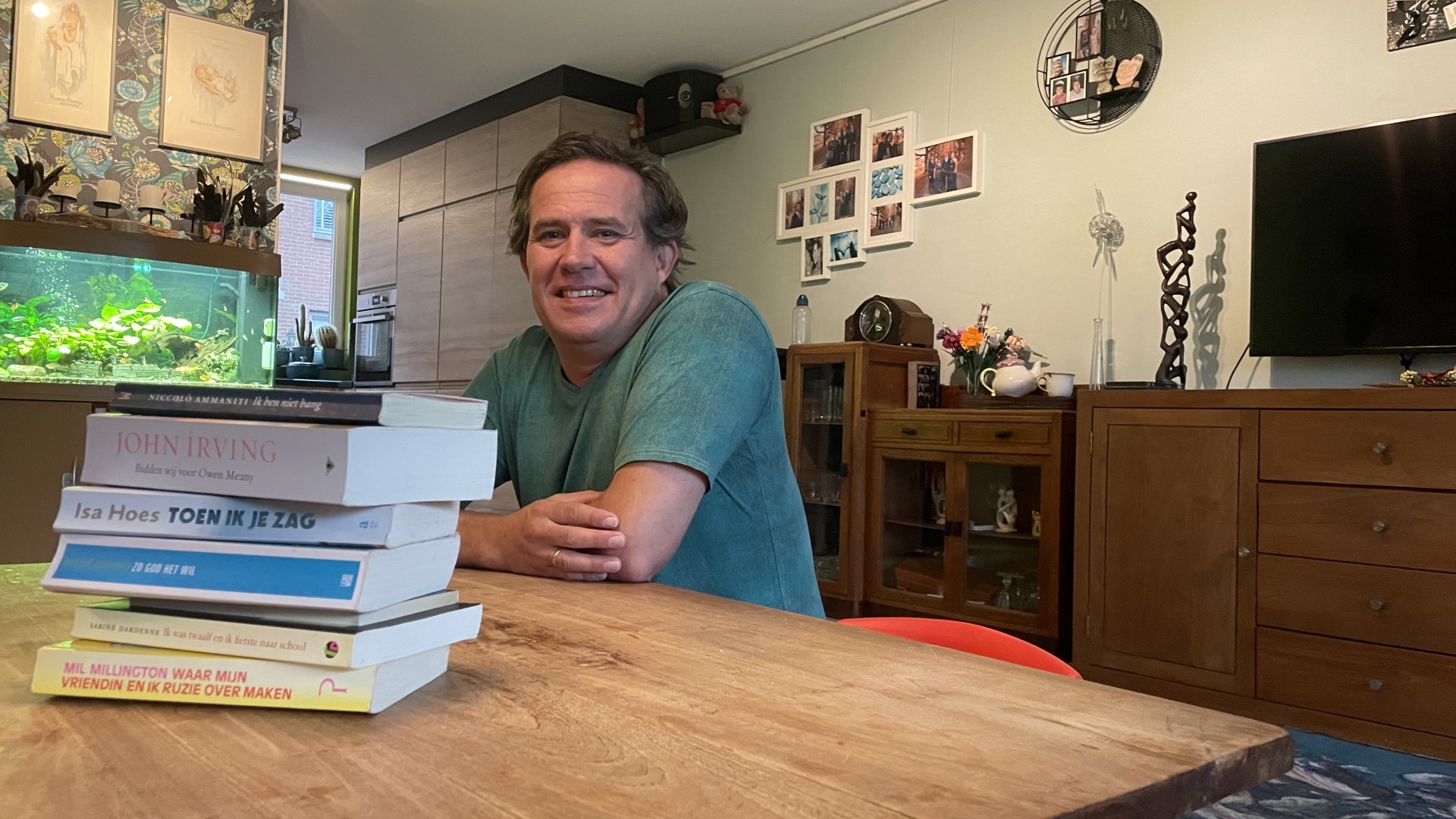 Man met groene trui zit aan een tafel met een stapel boeken in een woonkamer met een groot aquarium, houten meubels en fotolijsten aan de muur.