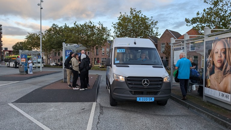 Bushalte met personen in gesprek en een geparkeerd Connexxion-busje met kenteken X-320-SK.