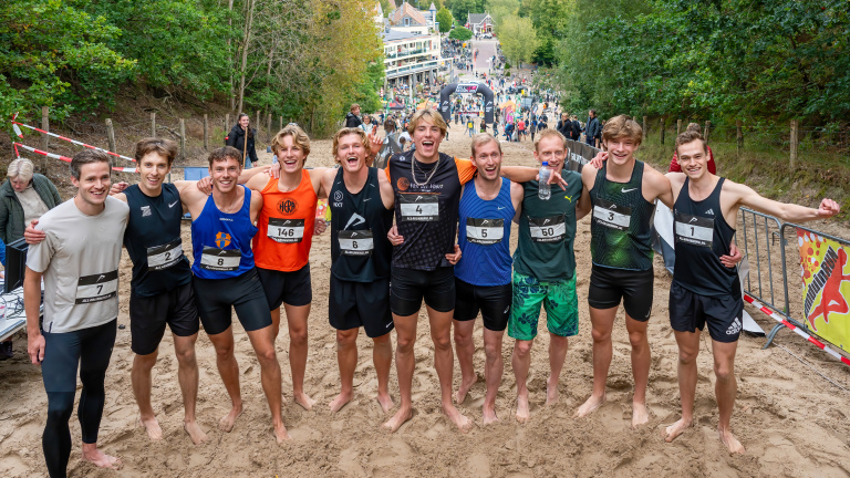 Een groep hardlopers poseert voor de camera na een race, omringd door bos en met een menigte op de achtergrond.