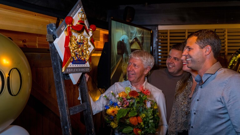 Man met bloemen wordt gefeliciteerd door drie mensen voor een schild met opschrift "Hofleverancier".