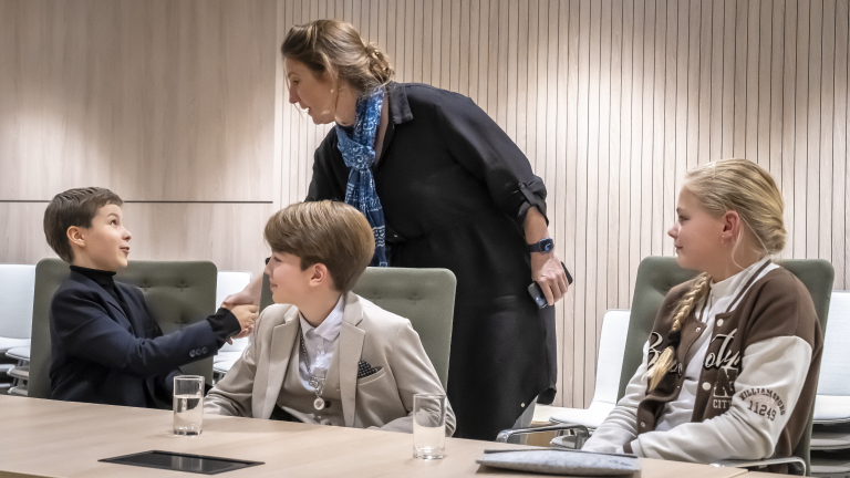 Vier kinderen zitten aan tafel in een vergaderzaal, terwijl een vrouw met een blauw sjaal over hen heen buigt en met hen praat.