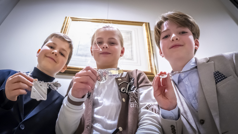 Kinderen houden medailles vast en poseren voor de foto, terwijl ze naar beneden kijken in de camera. Ze dragen nette kleding.