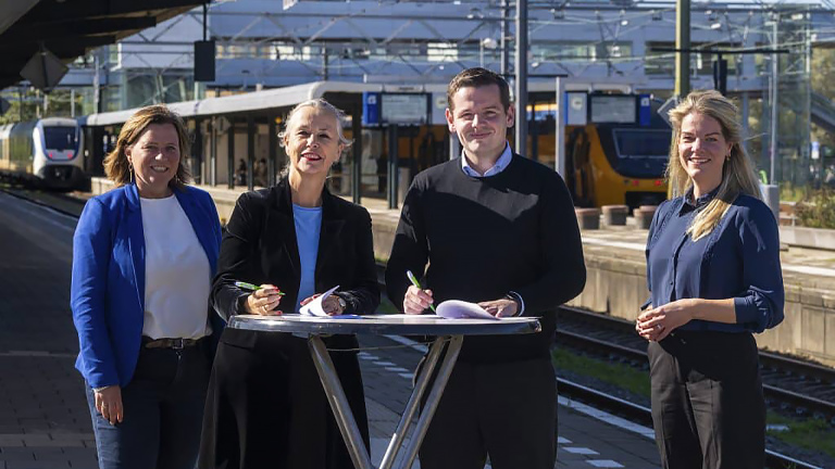 Vier mensen staan bij een tafel op een treinstation, met treinen op de achtergrond.