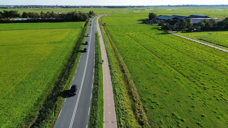 Luchtfoto van een landelijke weg met auto's en een fietspad tussen groene weilanden en een boerderij in de verte.