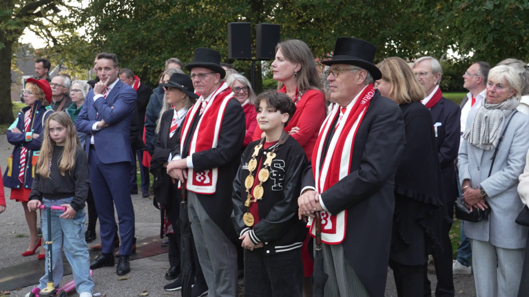 Een groep mensen, waaronder kinderen en volwassenen, sommigen gekleed in formele kleding en rood-witte sjaals, verzamelden zich buiten.