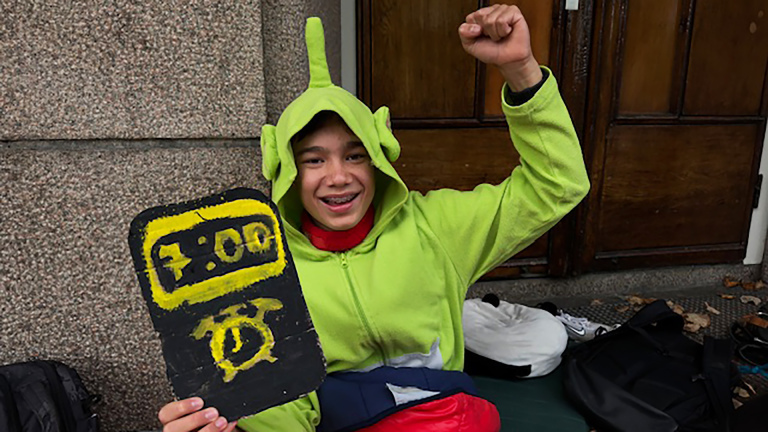 Jonge persoon in groene outfit met capuchon houdt een bord vast met een afbeelding van een klok en steekt zijn vuist in de lucht.