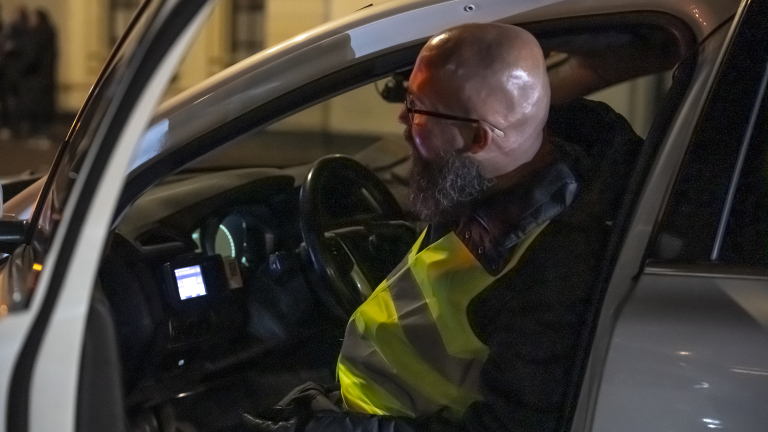 Man met een baard en bril draagt een fluorescerend geel vest in een auto.