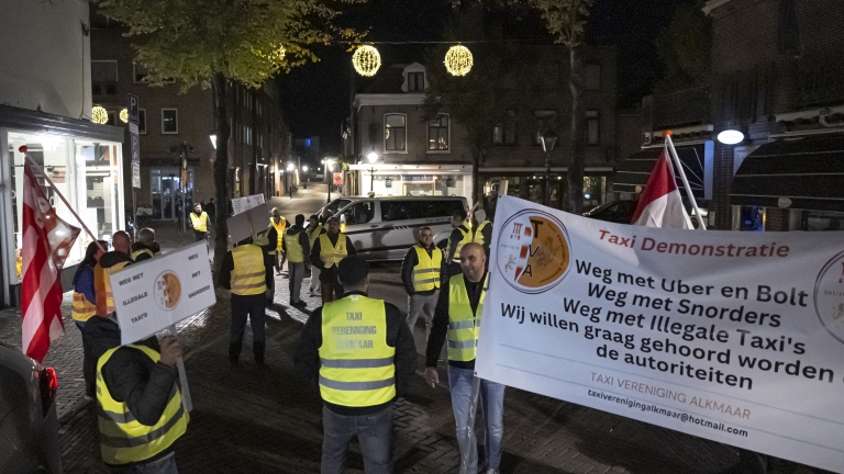 Mensen in gele hesjes protesteren in de avond op straat, met borden en een spandoek met teksten tegen Uber, Bolt en illegale taxi's.