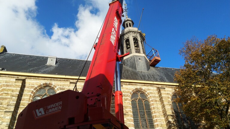 Een grote rode kraan van Winder Limmen staat voor een kerk met een hoge toren, onder een blauwe hemel met enkele wolken.