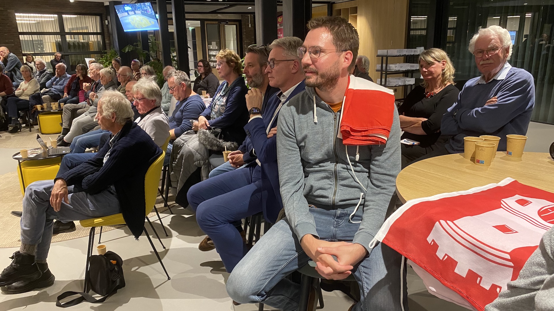 Groep mensen zit in een zaal, aandachtig kijkend naar een presentatie of scherm. Een man op de voorgrond heeft een rood-witte vlag op zijn schouder.