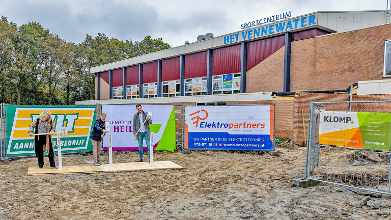 Drie personen poseren met scheppen op een bouwplaats voor een sportcentrum met verschillende bedrijfsbanners op de achtergrond.