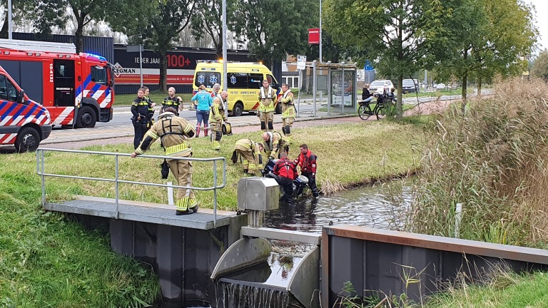 Brandweerlieden en hulpverleners helpen bij een waterreddingsactie langs een sloot, met een ambulance en brandweerwagen op de achtergrond.