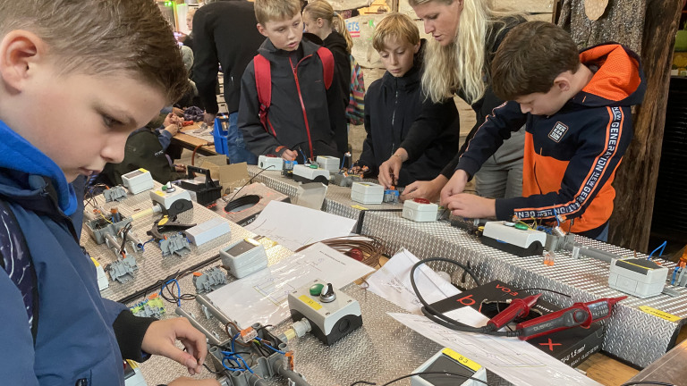 Kinderen werken aan een elektronica-project aan een tafel met schakelaars en bedrading, begeleid door een volwassene.