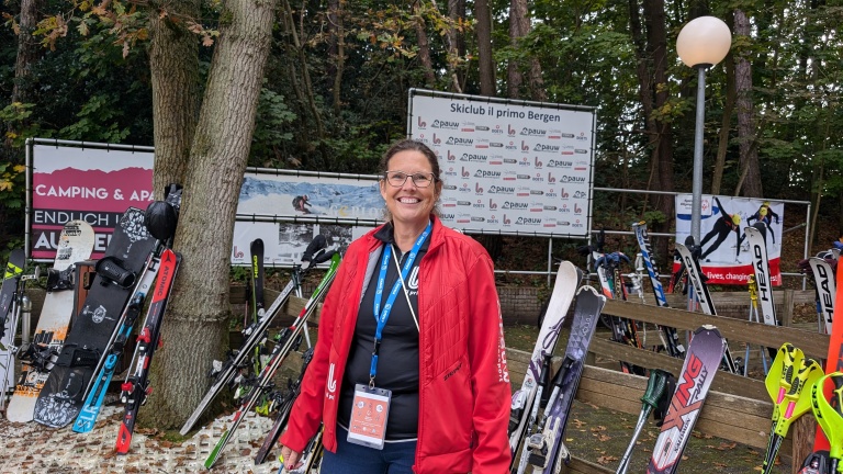 Vrouw in rode jas staat voor een verzameling ski's en snowboarden bij een skiclub.