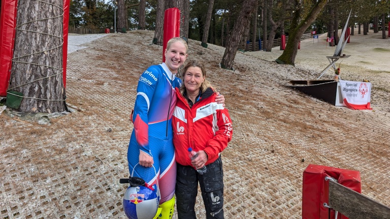 Twee personen poseren lachend voor de camera op een skibaan met een Special Olympics Nederland banner.