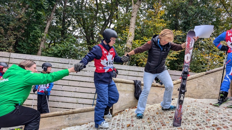 Mensen helpen elkaar terwijl ze zich voorbereiden op een buitenactiviteit met ski's op een helling met kunstmatige sneeuw, omringd door bomen.