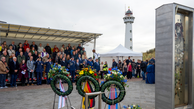 Mensen staan tijdens een herdenkingsplechtigheid bij een vuurtoren met kransen aan de voorgrond en een podium met toeschouwers.