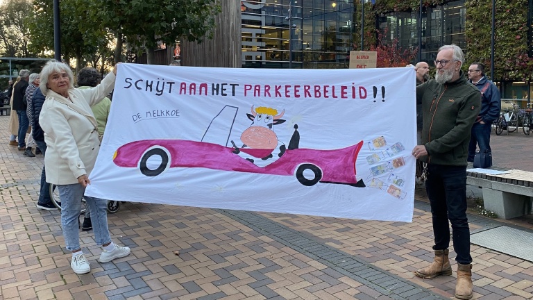 Twee mensen houden een spandoek met een cartoon van een koe in een roze auto vast, met de tekst "Schijt aan het parkeerbeleid!!".