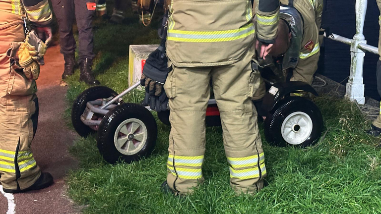 Brandweerlieden in uniform werken samen buiten op een grasveld naast een pad, met een klein voertuig met rubberen wielen.