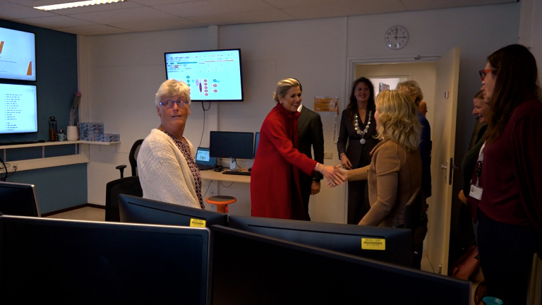 Een groep mensen in een kantooromgeving, een vrouw in een rode jas schudt handen met iemand. Schermen en bureaustoelen zijn zichtbaar.