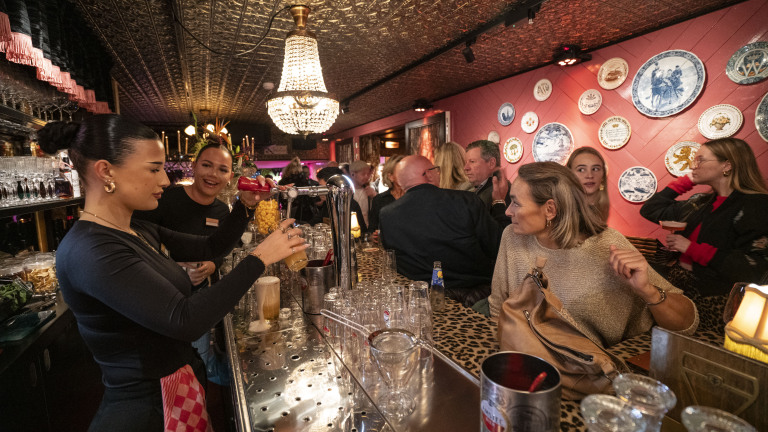 Bar met mensen, serveerster tapt bier achter een versierde toog, muren hangen vol borden.