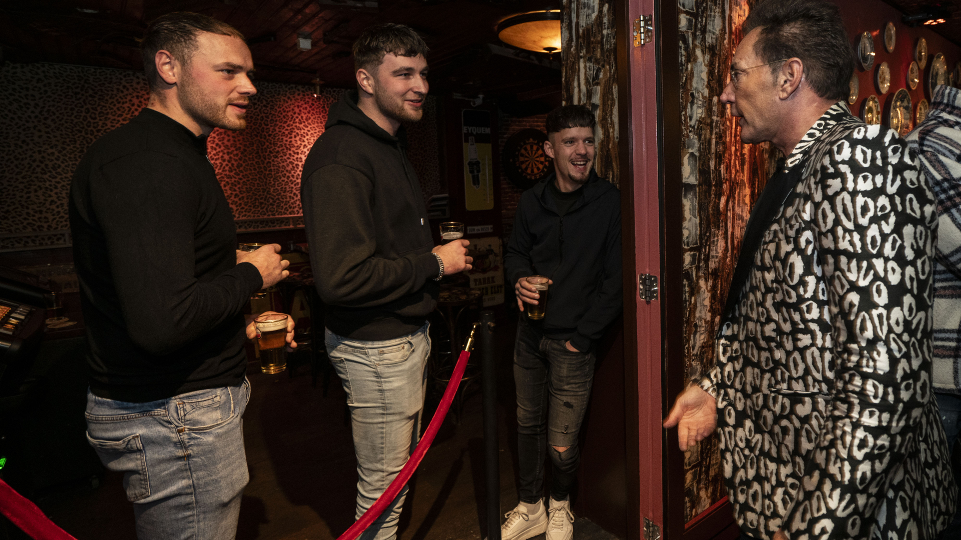 Vier mannen, drie met drankjes, kletsen in een bar. Een man draagt een opvallend zwart-wit luipaardpatroon jasje.