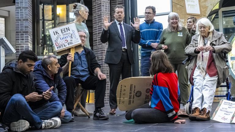 Een groep mensen, waarvan sommigen zitten en anderen staan, met protestborden tijdens een demonstratie in een binnenruimte. Eén bord met tekst "Ik ben heel erg boos".