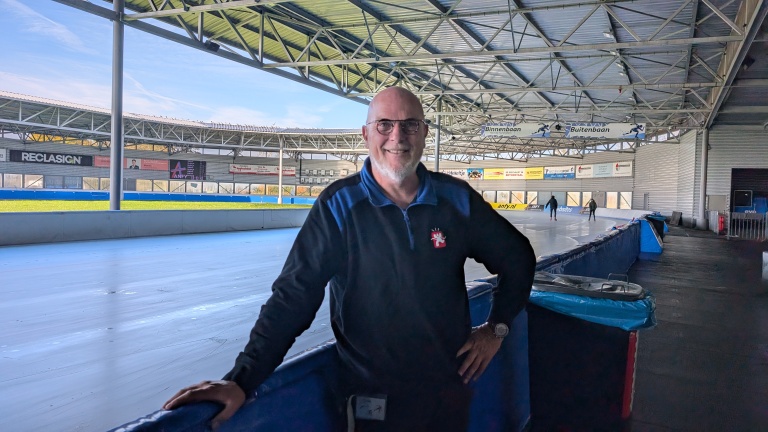 Man met bril en baard staat naast een ijsbaan in een overdekte schaatshal.