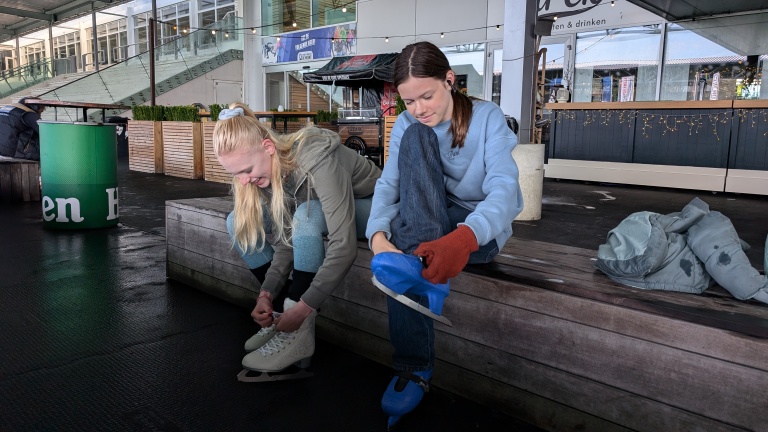 Twee vrouwen doen hun schaatsen aan op een bankje in een indoor skatecentrum.