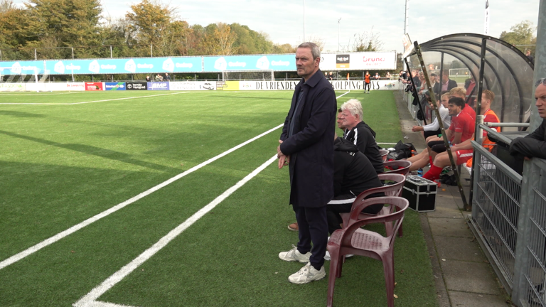 Man in donkere jas staat langs de zijlijn bij een voetbalveld, met mensen zittend op stoelen op de achtergrond.