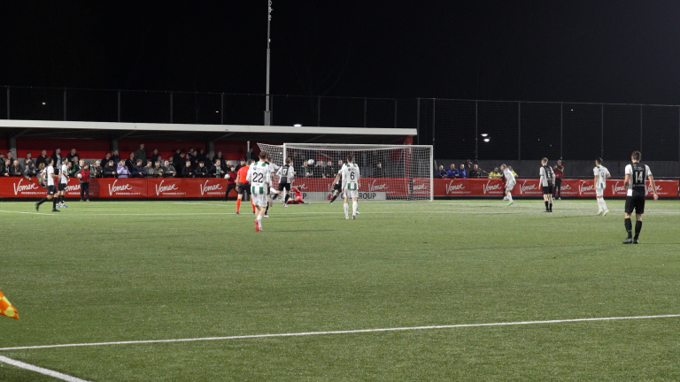 Voetbalwedstrijd op een verlicht veld met spelers in groene en zwart-witte tenues, toeschouwers op de tribune en een doelgebied in beeld.