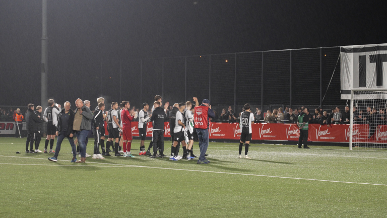 Voetballers en begeleiders op een veld na een avondwedstrijd, met toeschouwers op de achtergrond.