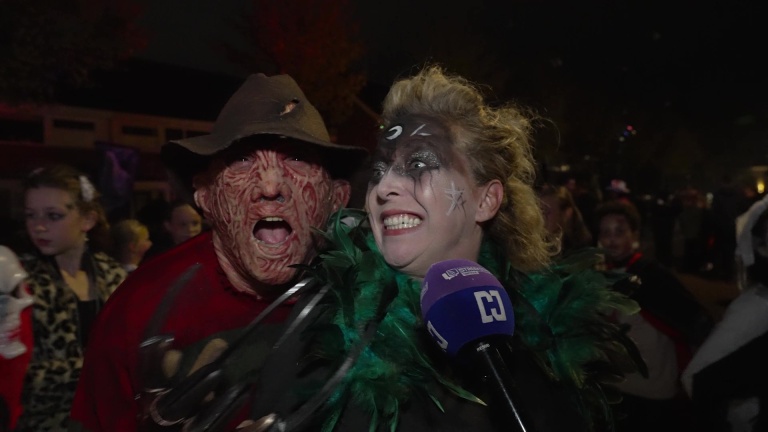 Twee mensen in Halloween-kostuums; de een draagt een verminkt gezichtsmasker en hoed, de ander heeft glinsterende make-up en een schminkpatroon. Beiden lachen met een microfoon in beeld.