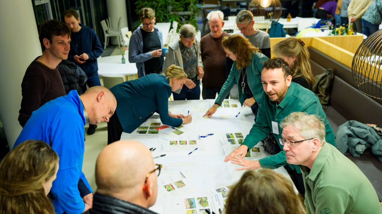 Groep mensen verzameld rond een tafel met papieren en pennen tijdens een vergadering of workshop in een binnenruimte.