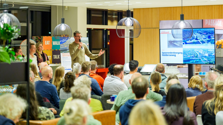 Een groep mensen luistert naar een presentatie in een kamer, met een spreker voor een scherm waarop "Spooronderdoorgang Helderseweg" staat.